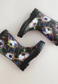 Black rain boots feature a soccer ball pattern with vibrant splashes of color. The sole is rugged and textured for grip.