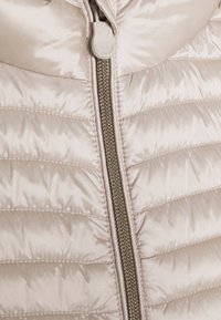 Jasnobeżowa kurtka puchowa z połyskującym wykończeniem, z poziomym pikowaniem, centralnym zamkiem błyskawicznym z suwakiem w złotym odcieniu oraz wysokim kołnierzem.