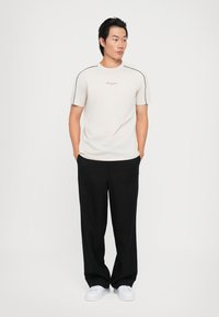 Camiseta de manga corta beige con detalles negros en los hombros, combinada con pantalones plisados negros y zapatillas blancas, sobre un fondo blanco liso.