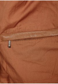 Tessuto marrone con una trama liscia presenta una tasca con zip nascosta con un tirante metallico e un accento in pelle sopra la cucitura della tasca.