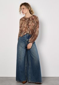 Braun gemusterte Bluse mit gerafftem Ausschnitt, kombiniert mit hochgeschnittenen, weit geschnittenen blauen Jeans. Schlichte, natürliche Stoffstruktur.