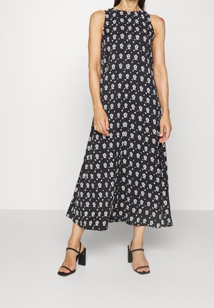 Femme portant une robe midi noire sans manches avec un petit imprimé floral blanc et des sandales noires à talons hauts, debout devant un fond uni.