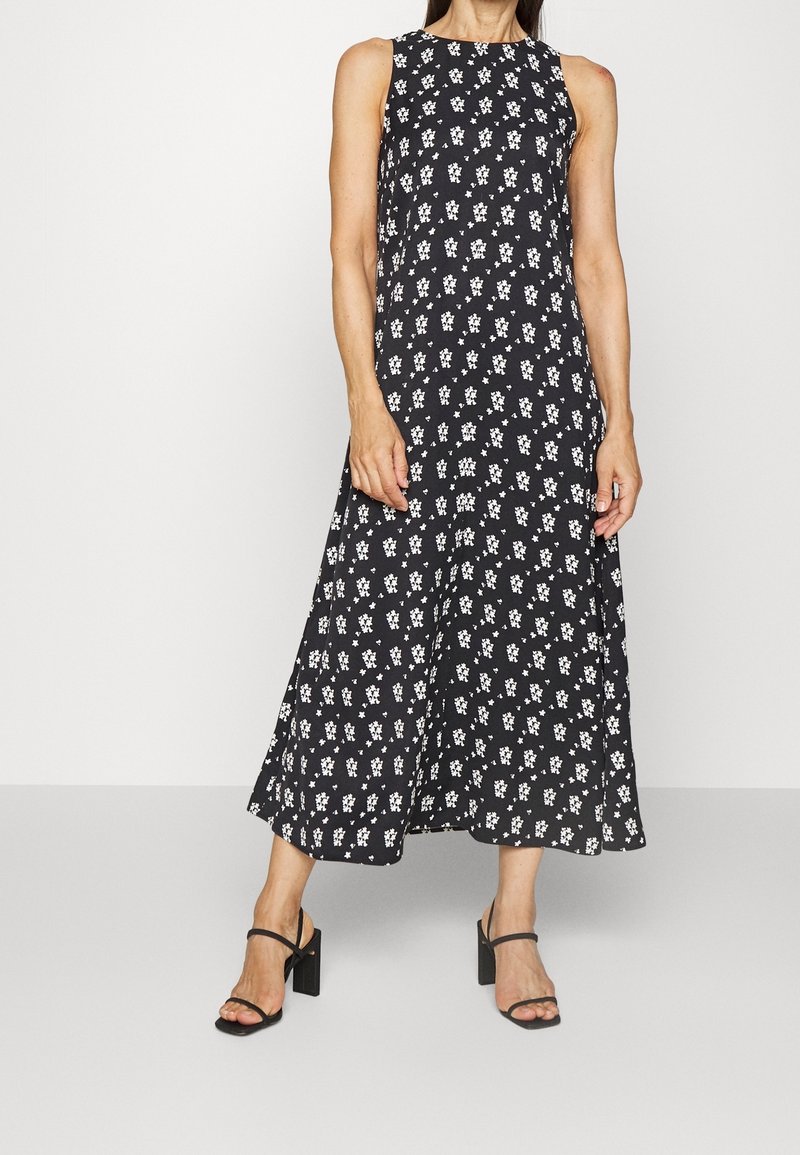Femme portant une robe midi noire sans manches avec un petit imprimé floral blanc et des sandales noires à talons hauts, debout devant un fond uni.