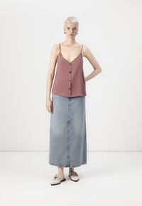 Roze camisole met knoopdetails, gecombineerd met een lange, lichtblauwe denimrok. Het model draagt neutrale slippers en heeft kort haar.