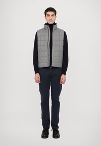 Gilet grigio imbottito con colletto alto, indossato sopra un dolcevita nero. Il modello indossa pantaloni scuri e scarpe nere su uno sfondo semplice.