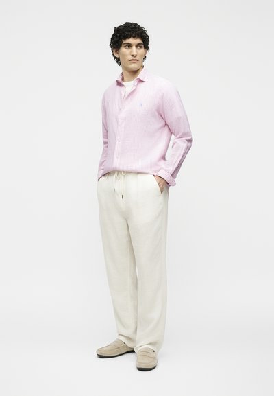 Άνδρας με σγουρά μαλλιά που φοράει ανοιχτό ροζ πουκάμισο με κουμπιά, λευκό παντελόνι με κορδόνι και μπεζ μοκασίνια, στέκεται με τα χέρια στις τσέπες.