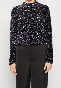Top nero con paillettes, maniche lunghe e colletto alto. Le paillettes presentano accenti viola, creando una superficie testurizzata e scintillante.