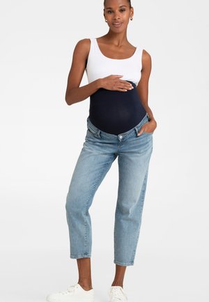 Gravid kvinde iført en hvid tanktop og blå barselsjeans holder på sin mave og står mod en ensfarvet baggrund.
