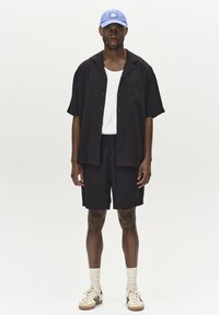 Kurzärmeliges schwarzes Hemd mit Knöpfen, weißes Tanktop, schwarze Shorts, blaue Mütze, cremefarbene Socken und schwarze Sneakers mit weißen Akzenten.