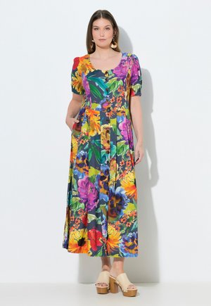 Vrouw die een kleurrijke bloemenmidi-jurk met knopen draagt, met pofmouwen en beige sandalen met plateauzool, staand tegen een witte achtergrond.