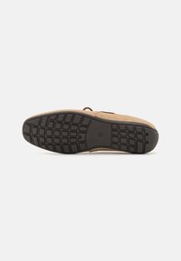 Tan shoe with a textured upper, brown laces, and a black rubber sole featuring a grid pattern for traction. Flat shape and casual design.