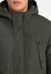 Olijfgroene, gewatteerde jas met een ritssluiting aan de voorkant, verstelbare capuchon en twee borstzakken met zwarte drukknoopsluitingen. Soepele textuur met subtiel patroon.