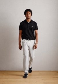 Zwart polo shirt, lichtgrijze chino's en zwarte sneakers. Model loopt met een gestructureerde achtergrond en een houten vloer. Eenvoudig ontwerp, neutrale kleuren.