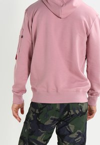 Sudadera con capucha rosa, con un bolsillo tipo canguro, puños y dobladillo acanalados, combinada con pantalones cargo verdes de camuflaje que cuentan con bolsillos laterales.