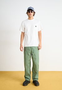 White cotton t-shirt with a small heart patch, green cotton pants, gray cap, and black chunky shoes, against a light backdrop.
