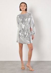 Robe en sequins argentés avec des manches longues, décolleté rond et ceinture à la taille. Texture lisse avec une surface brillante et réfléchissante.
