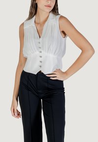Blusa bianca senza maniche con pieghe verticali, colletto e sei bottoni decorativi. Indossata con pantaloni neri aderenti con cuciture frontali.