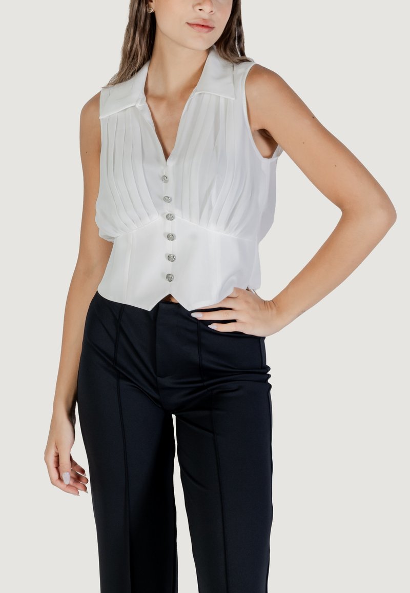 Blusa bianca senza maniche con pieghe verticali, colletto e sei bottoni decorativi. Indossata con pantaloni neri aderenti con cuciture frontali.