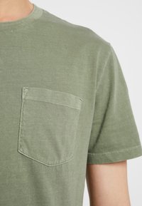 Gros plan sur une personne portant un t-shirt à manches courtes de couleur vert olive avec une poche poitrine sur le côté gauche.