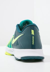 Zapatilla de entrenamiento de Nike en teal oscuro y amarillo neón, con parte superior de malla, collar acolchado y suela texturizada para tracción.