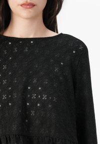Top in pizzo nero con un motivo geometrico, tessuto testurizzato, scollatura ampia e orlo inferiore leggermente arricciato.