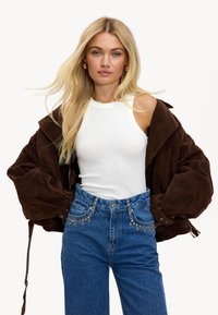 Veste oversize en daim marron, débardeur en coton blanc à côtes et jeans taille haute bleu avec des clous argentés le long des poches.