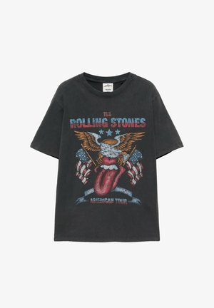 Mladý muž má na sebe vyblednuté čierne tričko Rolling Stones, hnedé kraťasy po kolená, biele ponožky a hnedé mokasíny a stojí opretý o bielu stenu.