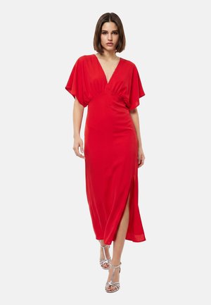 Giovane donna con capelli castani corti che indossa un vestito midi rosso con scollo a V, maniche corte e spacco frontale, cammina con sandali argentati con tacco alto.