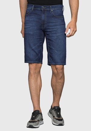 Uomo che indossa pantaloncini di jeans blu scuro e scarpe da ginnastica grigio nere, in piedi davanti a uno sfondo chiaro e semplice.