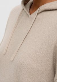 Beżowa bluza z kapturem wykonana z miękkiej dzianiny. Posiada przednią kieszeń typu kangurka i sznurki ściągające. Gładka tekstura i luźny fason.