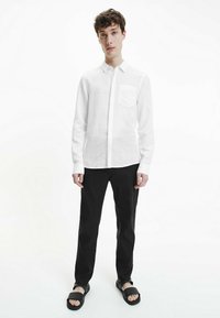 Camicia bianca a maniche lunghe in lino con taschino sul petto, abbinata a pantaloni neri e sandali scuri senza lacci. Il modello è in piedi davanti a uno sfondo bianco uniforme.