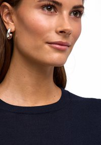 Granatowy sweter z dzianiny z prążkowanym dekoltem; modelka nosi duże srebrne kolczyki koła; gładka tekstura skóry, lekki makijaż i brązowe włosy.