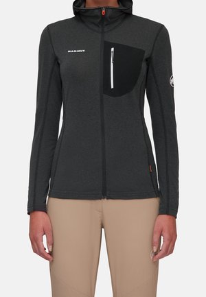 Ciemnoszara kurtka z pełnym zamkiem błyskawicznym, czarna zapinana na zamek kieszeń na piersi, logo Marmot na lewej piersi oraz beżowe spodnie na stojącej osobie.