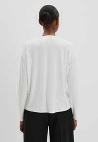 Haut blanc à manches longues avec une coupe décontractée, texture lisse et ourlet arrondi, associé à un pantalon noir. Design minimal sans motifs visibles.