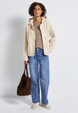 Femme portant une veste polaire à capuche crème, un haut beige, un jean bleu à jambes larges, des baskets blanches, tenant un grand sac fourre-tout en daim marron.