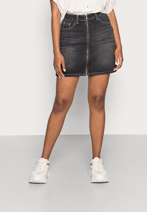 Vrouw draagt een zwarte denim minirok met zichtbare ritssluiting aan de voorkant, wit overhemd ingestopt en witte chunky sneakers, staand op een grijze vloer.