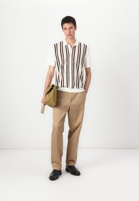 Srajca s kratkimi rokavi in ovratnikom z vertikalnimi rjavimi in črnimi črtami, v kombinaciji s svetlo bež hlačami in črnimi gležnjarji. Drži olivnozeleno torbo.