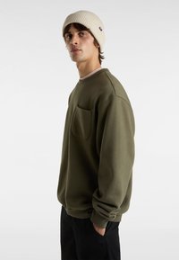 Olijfgroen sweatshirt met een relaxte pasvorm, geribde ronde hals, lange mouwen en een voorzak. Draag het met een beige beanie.