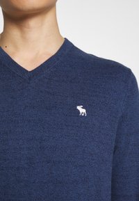 Marineblauwe V-hals trui van textuurgebreide stof, met een klein wit elandlogo op de linkerkant van de borst.