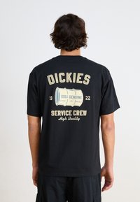 T-shirt en coton noir avec un grand imprimé dans le dos, comportant du texte beige et un design en forme de baril. Col rond et manches courtes.