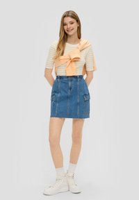 Jeansrock mit rohem Saum, ausgestattet mit zwei Fronttaschen. Kombiniert mit einem gestreiften Shirt in Weiß und Gelb sowie einem hellorangefarbenen Pullover, der um die Taille gebunden ist.