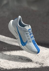Svetlo sivi in modri Nike tekaški čevlji z belimi vezalkami, ki lebdi nad asfaltom z belo pobarvano črto pod njimi.