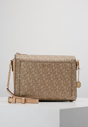 Beige crossbody taske med DKNY logo mønster, guldkæde rem, frontlomme og gyldent cirkulært vedhæng på en hvid overflade.