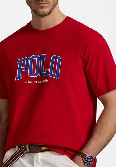 Red cotton t-shirt featuring large white and blue "POLO" text with "Ralph Lauren" below. Smooth texture, short sleeves, standard fit.