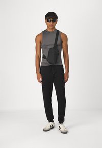 Sac bandoulière noir avec sangle réglable, compartiments zippés, fabriqué en tissu durable. Le modèle porte un haut sans manches gris et un pantalon noir.