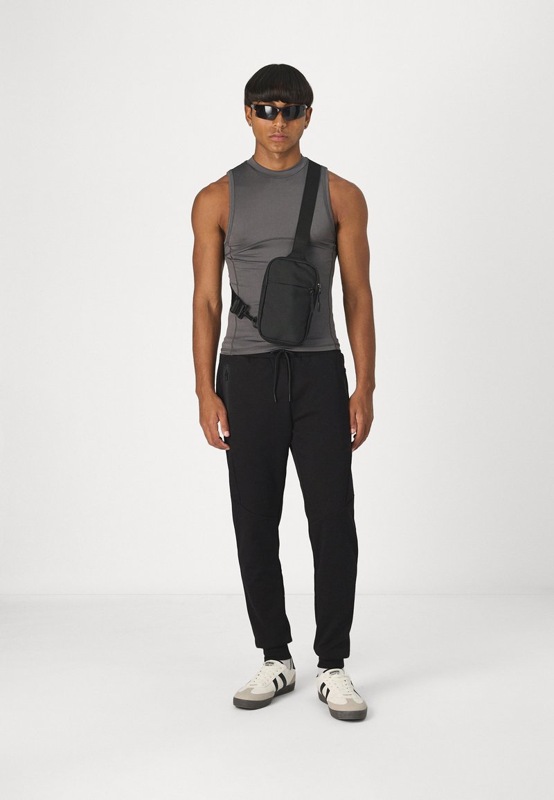 Sac bandoulière noir avec sangle réglable, compartiments zippés, fabriqué en tissu durable. Le modèle porte un haut sans manches gris et un pantalon noir.