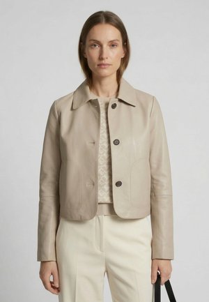 Woman wearing a beige cropped leather jacket over a patterned sweater and cream trousers, holding a black handbag by her side.