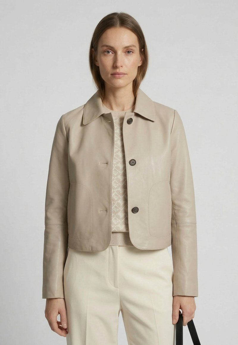 Frau trägt eine beige kurze Lederjacke über einem gemusterten Pullover und cremefarbenen Hosen, hält eine schwarze Handtasche an ihrer Seite.