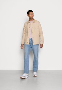 Casaco de algodão bege com dois bolsos frontais, t-shirt lilás clara por baixo, combinado com jeans azuis de perna reta e sapatilhas brancas.