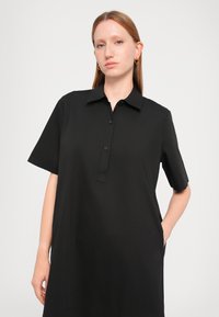 Black short-sleeve shirt dress made of smooth fabric, featuring a buttoned placket, pointed collar, and side pockets.